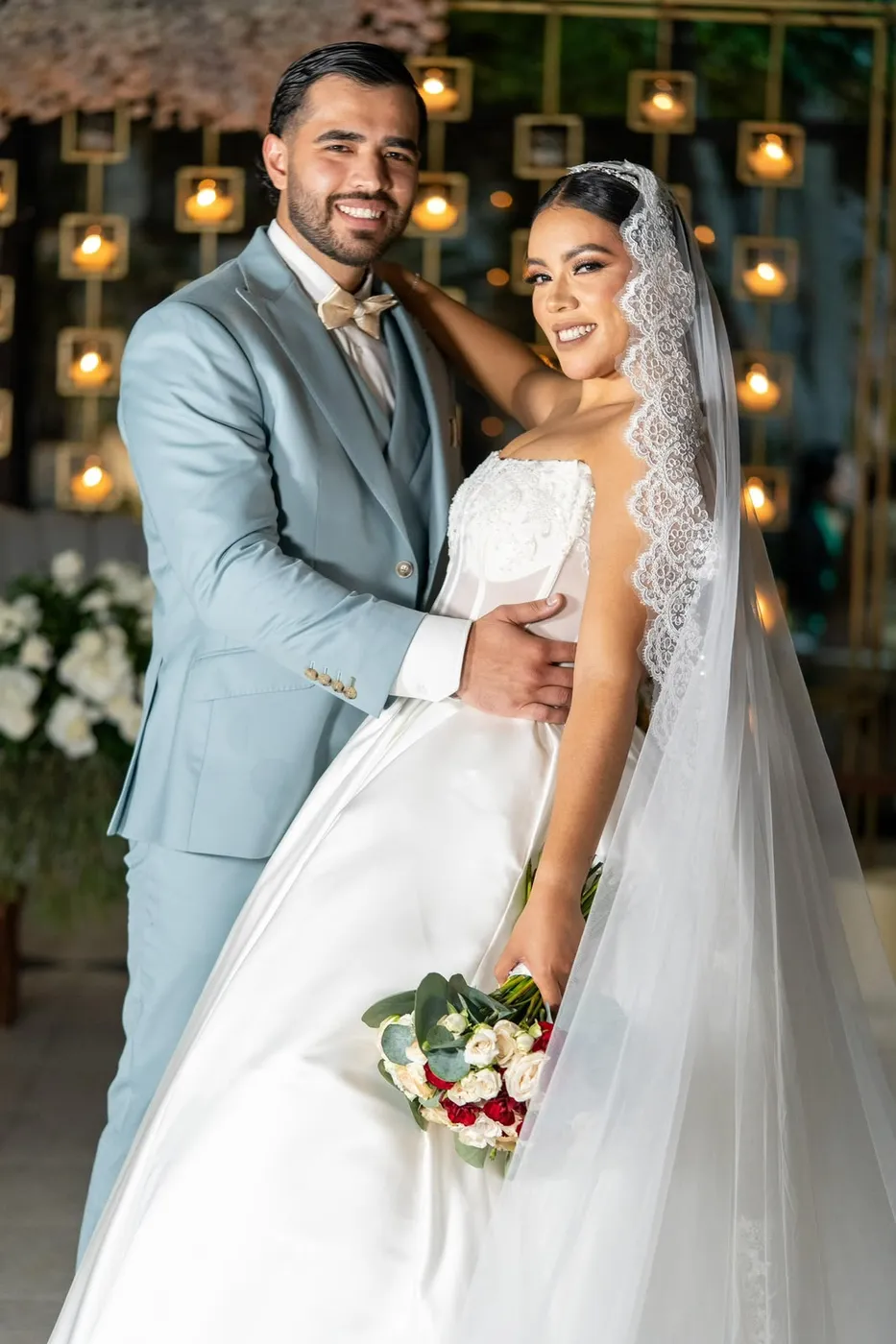 Novia con velo mantilla de encaje junto a su pareja, pared de luces doradas
