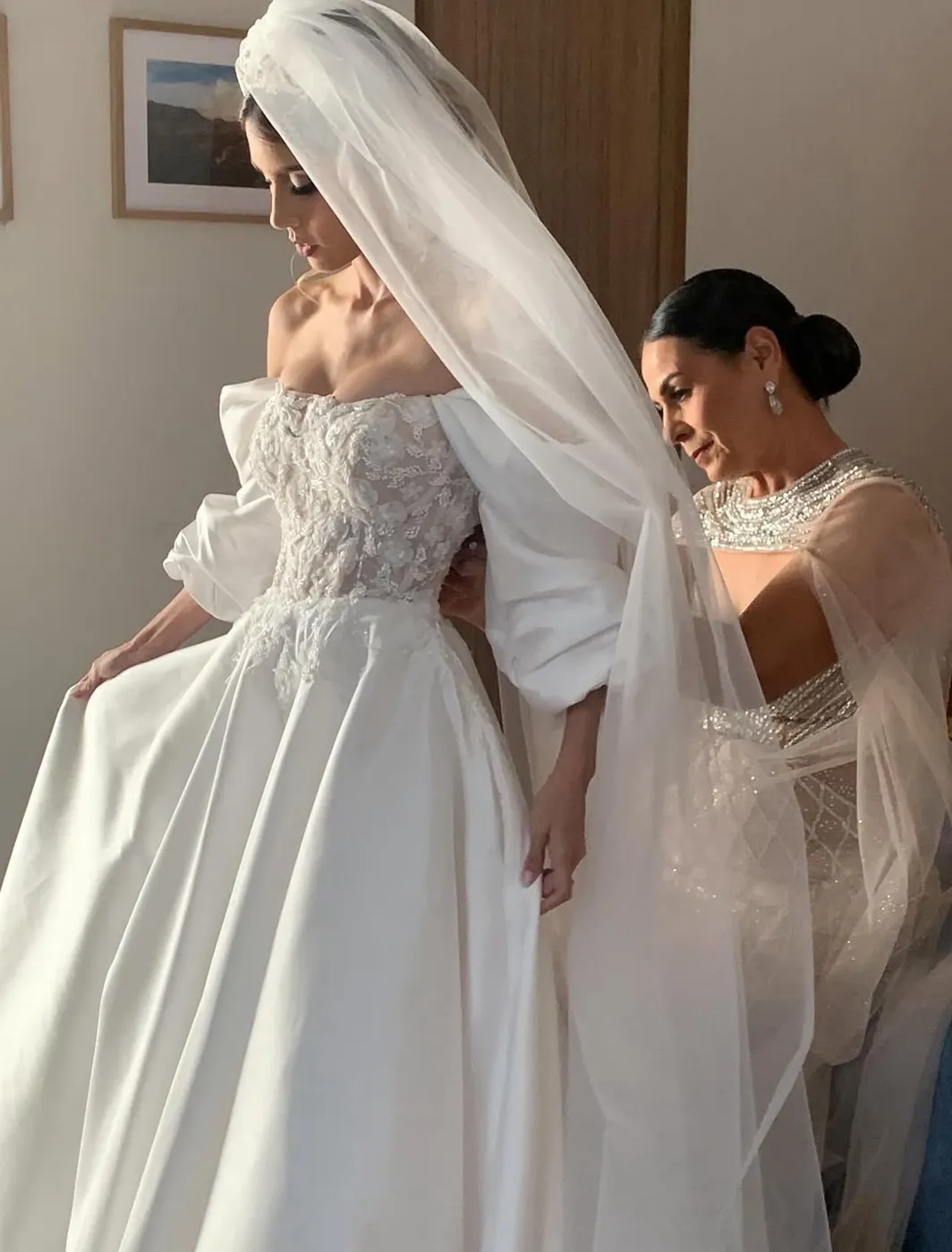 Novia de perfil con velo mientras le abrochan el vestido de pedrería